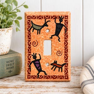 May include: A rectangular light switch cover plate with a speckled orange finish. The plate features a dark red geometric border and black line art illustrations of two stylized human figures and two animals, with two screw holes and a centre cutout.