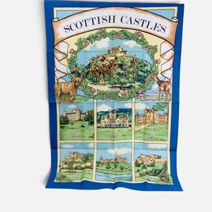 Puede incluir: Un pa&ntilde;o de cocina rectangular con borde azul que presenta el texto SCOTTISH CASTLES en la parte superior. El dise&ntilde;o incluye una ilustraci&oacute;n central de un castillo rodeada por una corona floral, una vaca Highland, un ciervo y seis escenas.