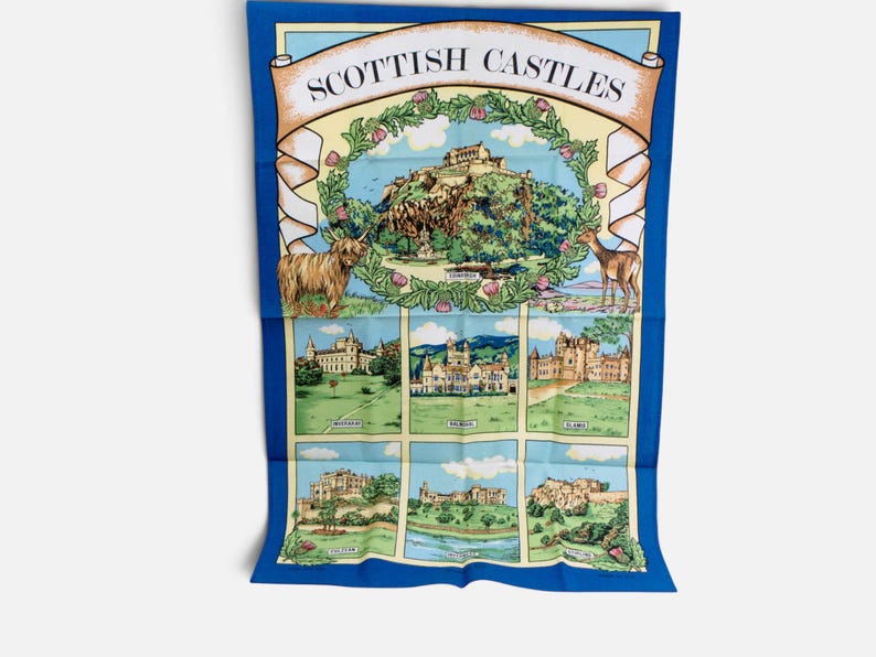 Puede incluir: Un pa&ntilde;o de cocina rectangular con borde azul que presenta el texto SCOTTISH CASTLES en la parte superior. El dise&ntilde;o incluye una ilustraci&oacute;n central de un castillo rodeada por una corona floral, una vaca Highland, un ciervo y seis escenas.