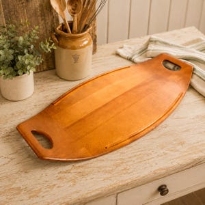 May include: An oval-shaped wooden serving tray with a smooth, polished finish and two integrated cutout handles on the ends. The tray features a warm, light brown wood grain pattern and a slightly raised edge along the sides for containment.