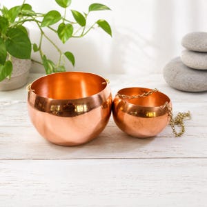 May include: Two polished copper bowls with a rounded shape. The larger bowl is plain, while the smaller bowl features a thin gold-toned metal chain attached to its sides for hanging. Both bowls have a smooth, reflective surface and open tops.