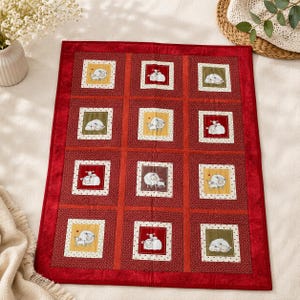 May include: A rectangular quilted wall hanging featuring a grid of twelve square panels. Each panel displays a cartoon illustration of sheep on backgrounds of yellow, green, or red. The quilt is bordered by a solid red fabric frame.