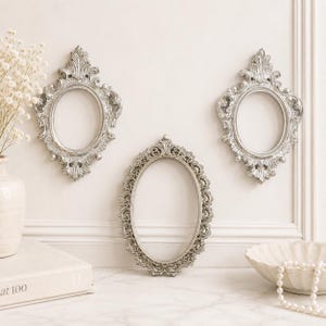 May include: Three oval picture frames with ornate, silver-coloured detailing. Two frames are positioned higher on the wall with elaborate scrollwork at the top and bottom, while the third frame is centred below with a detailed, textured border.