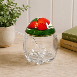 May include: A clear glass jar with a rounded base and three small feet, featuring a lid decorated with a vibrant red strawberry and green leaf design. A small, clear glass spoon rests inside the jar, which has a textured, star-like pattern on its bottom surface.
