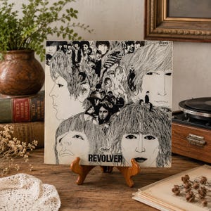 Puede incluir: La portada del disco de vinilo Revolver de The Beatles presenta un collage de dibujos en blanco y negro de los rostros y el cabello de los miembros de la banda. El logotipo de Capitol Records y el título REVOLVER están impresos en negro.