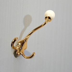 May include: A brass wall hook with a double hook design and a curved arm. The hook features a white, spherical knob at the end of the arm. The hook is mounted on a light blue wall.