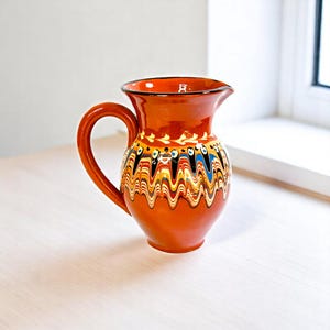Peut inclure: Une cruche en céramique avec une anse, peinte dans des tons orange, marron, jaune, bleu et noir. La cruche présente un motif ondulé décoratif sur le corps et un motif floral simple près du bord. Poterie de style traditionnel.