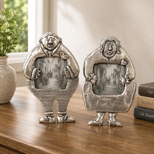 May include: Two silver-toned metal picture frames shaped like stylized figures. The left frame depicts a figure in striped trousers, while the right frame shows a figure in a dress. Both frames feature a central rectangular opening for photos.