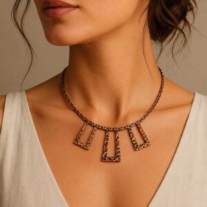May include: A bronze-coloured necklace featuring a chain and three pendant sections. Each pendant has a rectangular shape with a textured surface. The necklace is worn on a person's neck, against a neutral background.