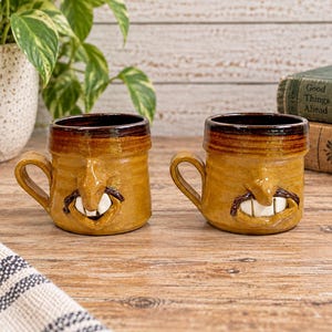 May include: Two matching ceramic mugs with a glossy mustard-yellow glaze and a dark brown rim. Each mug features a sculpted human-like nose, a dark brown moustache, and two prominent white teeth protruding from the mouth area, with a side handle.