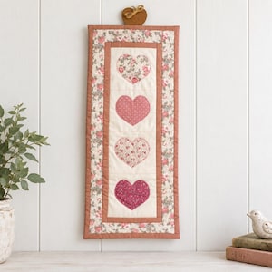Peut inclure: Une tenture murale rectangulaire verticale en tissu présentant quatre cœurs appliqués en colonne. Les cœurs sont disposés sur un fond crème avec une bordure florale dans des tons de rose, vert et marron. Un petit cœur en bois est fixé en haut.