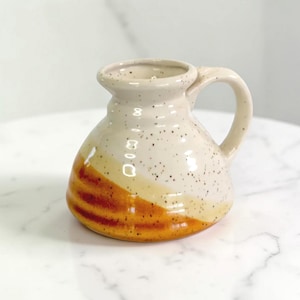 May include: A handmade ceramic jug with a handle. The jug has a cream-coloured body with speckled detailing and a band of orange and yellow glaze. The jug is designed with a wide base and a narrow opening.