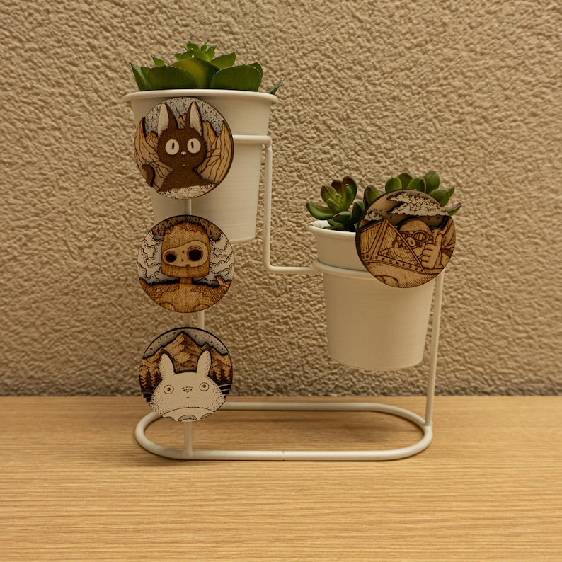 Set of four round wooden magnets inspired by Studio Ghibli characters — Totoro, Porco Rosso, Jiji, and the Laputa robot. Displayed on white plant pots with green succulents, showing their natural wood tones and engraved details. Handmade from layered plywood and MDF, each magnet measures 5 cm in diameter. Perfect for decoration or as a gift for anime and Studio Ghibli fans.