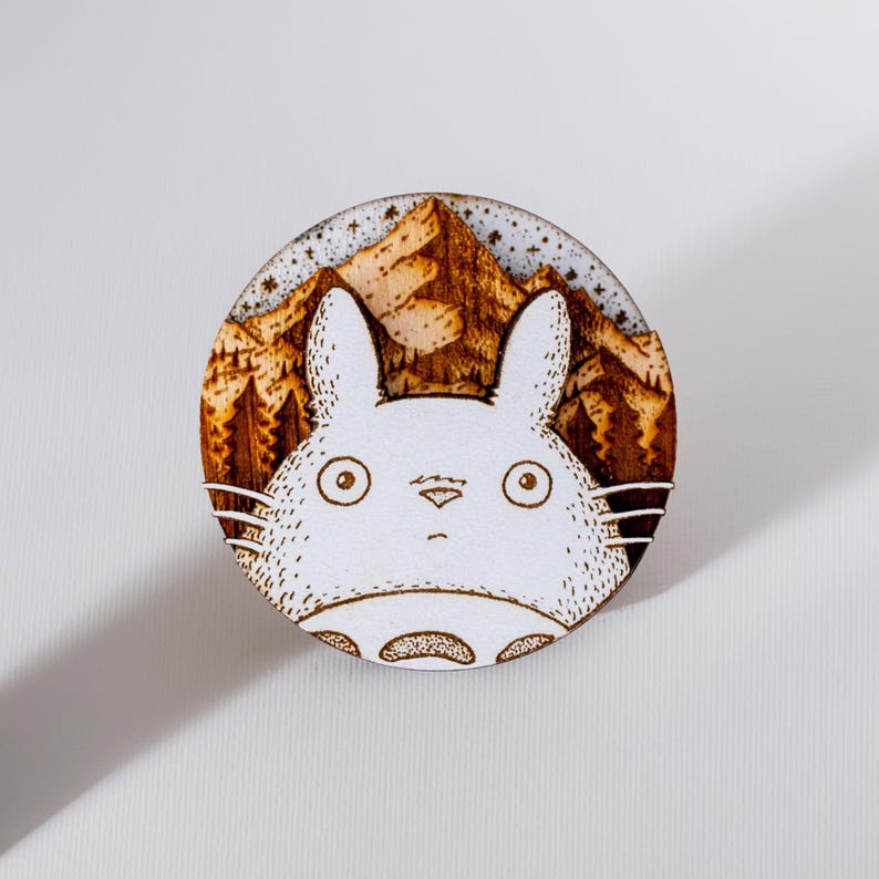 Round wooden magnet inspired by Totoro from My Neighbor Totoro. Handmade from layered plywood and MDF, it features a detailed engraving of Totoro’s face against a mountain and forest background in warm brown tones. Measures approximately 5 cm in diameter. A charming, nature-inspired piece perfect for Studio Ghibli or anime fans.