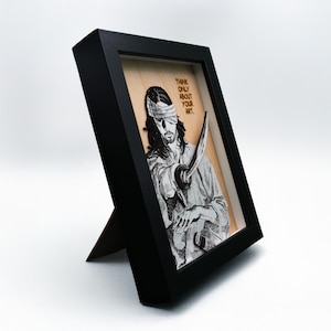 May include: A black framed wood wall art piece with a black and white image of a blindfolded man holding a sword. The text "Think only about your art." is written in gold lettering on the wood.