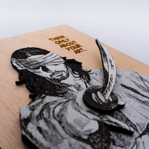 May include: A wooden wall art piece featuring a black and white illustration of a blindfolded man holding a sword. The text "Think only about your art." is engraved on the wood.