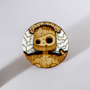Round wooden magnet inspired by the guardian robot from Castle in the Sky. Handmade from layered plywood and MDF, it features a detailed engraving of the robot surrounded by clouds in warm golden and brown tones. Measures approximately 5 cm in diameter. A unique handmade piece perfect for Studio Ghibli collectors and anime enthusiasts.