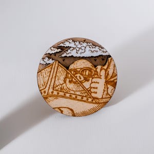 Round wooden magnet inspired by Porco Rosso from Porco Rosso. Handmade from layered plywood and MDF, it features a detailed engraving of the pilot in his airplane flying among clouds and mountains in warm brown tones. Measures approximately 5 cm in diameter. A unique handmade piece perfect for Studio Ghibli fans and anime lovers.