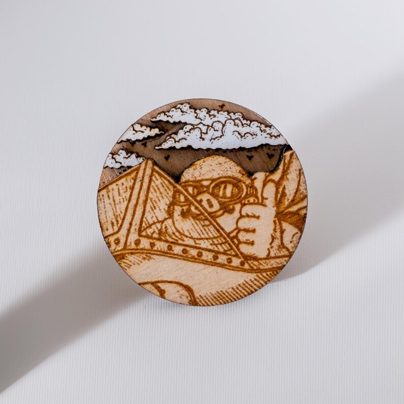 Round wooden magnet inspired by Porco Rosso from Porco Rosso. Handmade from layered plywood and MDF, it features a detailed engraving of the pilot in his airplane flying among clouds and mountains in warm brown tones. Measures approximately 5 cm in diameter. A unique handmade piece perfect for Studio Ghibli fans and anime lovers.