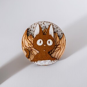 Round wooden magnet inspired by Jiji, the black cat from Kiki’s Delivery Service. Handmade from layered plywood and MDF, it features a detailed engraving of Jiji with large eyes against a mountain background in warm brown tones. Measures approximately 5 cm in diameter. A charming handmade piece ideal for Studio Ghibli fans and cat lovers alike.