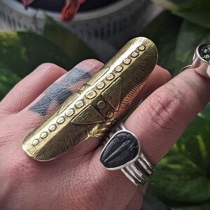 May include: A brass knuckle ring with a geometric design. The ring is worn on a finger with a tattoo.