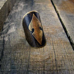 May include: A wide, brass ring with a textured, dark brown finish. The ring features a raised, geometric design in the center.