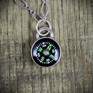 May include: A silver necklace with a compass pendant. The compass face is black with green lettering and markings. The compass is pointing north.