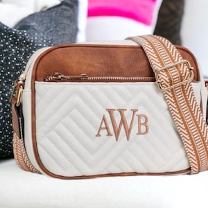 May include: A cream-colored crossbody bag with a brown top and strap. The bag features a quilted pattern and the embroidered initials "AWB". The adjustable strap has a geometric design.