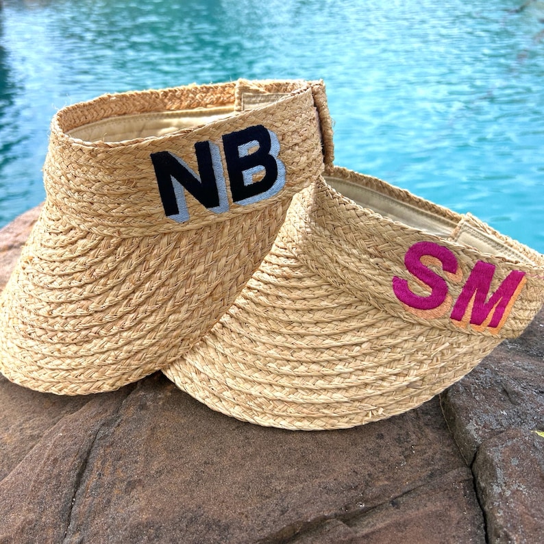 Peut inclure: Deux visi&egrave;res en paille tress&eacute;e. L'une porte les lettres "NB" brod&eacute;es en noir avec un contour bleu clair. L'autre porte les lettres "SM" brod&eacute;es en rose avec un contour orange. Les visi&egrave;res sont sur un rocher avec une piscine en arri&egrave;re-plan.