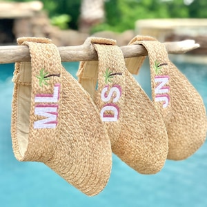 May include: Three woven straw visors with personalized initials embroidered in white and pink, each featuring a small palm tree design. The visors are hanging on a wooden branch against a blurred blue water background.