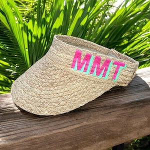 May include: A woven straw visor with the letters "MMT" in pink with a turquoise outline. The visor is sitting on a wooden surface with green foliage in the background. The visor is designed to shield the eyes from the sun.