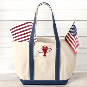 May include: Cream-colored tote bag with navy blue accents, featuring a red lobster design and initials. A red and white striped towel is inside, and an American flag is attached. The bag has navy blue handles and trim.