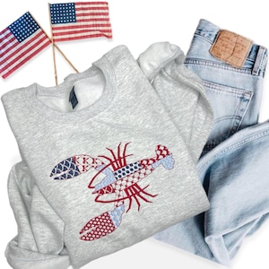 May include: A light gray sweatshirt featuring an embroidered lobster design in red, white, and blue patterns. The image includes a pair of light blue jeans and two small American flags, suggesting a patriotic theme.