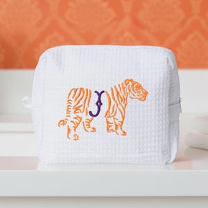 May include: White, square cosmetic bag with a waffle texture. An embroidered orange tiger with purple initial is on the front. The bag has a zipper closure and a small handle on the side. The background is orange.
