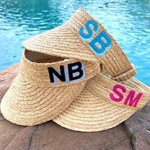May include: Three woven straw visors with embroidered initials. One visor has "SB" in light blue, another has "NB" in dark blue, and the third has "SM" in pink. The visors are displayed against a backdrop of water.