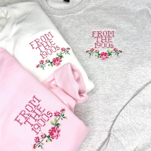May include: Three sweatshirts in white, pink, and gray, each embroidered with the text "FROM THE 1900s" in pink, accompanied by a floral design. The sweatshirts are folded, showcasing the embroidered details.