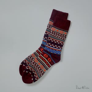 Op de afbeelding: Een paar bordeauxrode sokken met een kleurrijk geometrisch patroon. Het patroon bevat de kleuren oranje, blauw, wit en grijs.
