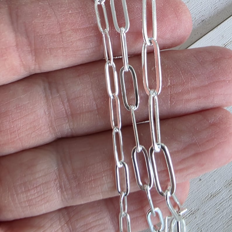 Pu&ograve; includere: Primo piano di tre fili di una catena d'argento con maglie ovali allungate. La catena &egrave; tenuta contro uno sfondo sfocato, mostrando la qualit&agrave; riflettente del metallo. Le maglie sono uniformi per dimensioni e forma, creando un'estetica elegante e moderna.
