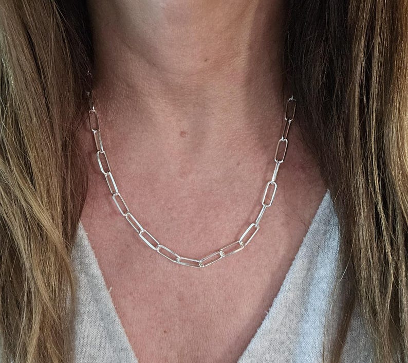 Pu&ograve; includere: Una collana a catena d'argento con maglie rettangolari allungate. La collana &egrave; indossata intorno al collo e le maglie sono visibili sulla pelle e sui vestiti. La catena &egrave; un gioiello semplice ed elegante.