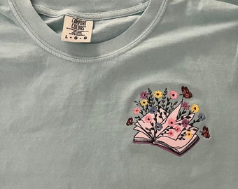 May include: A light blue t-shirt featuring an embroidered design of an open book with flowers and butterflies. The shirt has a tag that reads "Comfort Colors" and "L-G-G".
