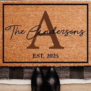 May include: A rectangular doormat with a brown coir surface and a black border. The mat features the text "The Cindersons" in a cursive font, a large letter "A", and "EST. 2025". The mat is placed on a black and white checkered rug.