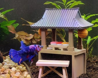 May include: A blue and purple betta fish swims near a miniature beige structure resembling a small pavilion with a gray roof, two orange lanterns, and a light pink bench. The structure features a small table holding three tiny colorful sushi pieces.