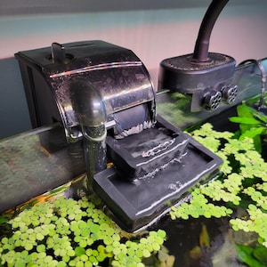 May include: A black aquarium filter with a water flow directed into a black plastic waterfall-style water feature. The filter is attached to the side of the aquarium. The aquarium is filled with water and has green plants growing in it.