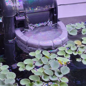 May include: A gray, semi-circular stone-textured aquarium filter baffle positioned beneath a water outlet. The structure features a brick-like pattern and is surrounded by floating green aquatic plants in a tank with dark water.