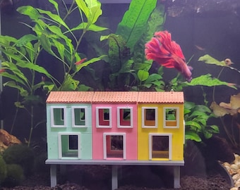 May include: A three-section aquarium ornament shaped like a row of houses with a light green, pink, and yellow facade, white window and door frames, and a textured orange roof, all supported by a gray platform with four legs inside a fish tank.