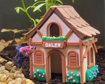 May include: A colorful betta fish swims near a miniature house aquarium decoration. The house is tan, brown, and white with the name "GALEN" above the door. Green and pink accents decorate the house.