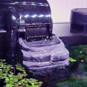 May include: A black aquarium filter with a waterfall feature. The filter is positioned above a gray, layered rock structure, with water cascading down. Green aquatic plants and floating leaves are visible in the water.
