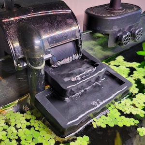 May include: A black aquarium hang-on-back power filter with a rectangular spillway tray. The device features a curved intake tube and a textured plastic housing, with water flowing over the spillway into a tank filled with small green floating plants.