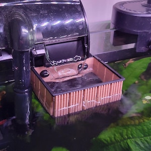 May include: A black aquarium filter with water flowing into a brown and black filter box. The filter box is submerged in water with green plants visible in the background. The filter is designed to clean the water in the aquarium.