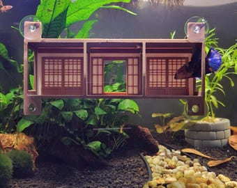 May include: A decorative aquarium ornament featuring a miniature Japanese-style house with three sections. The structure is brown with intricate door and window designs, and is held in place by suction cups. A blue and black betta fish swims nearby.